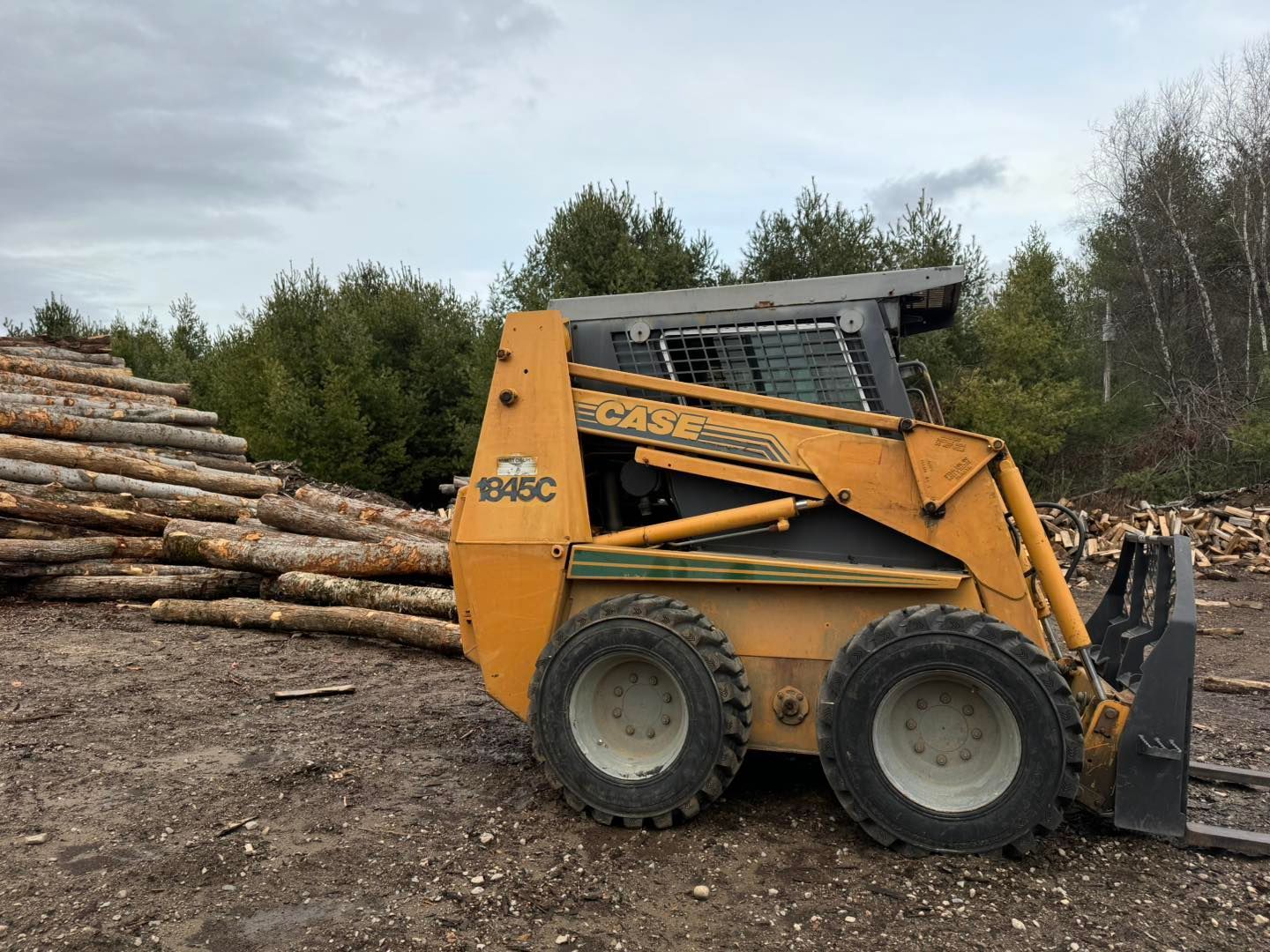 A yellow skid steer loader is parked in front of a pile of logs.