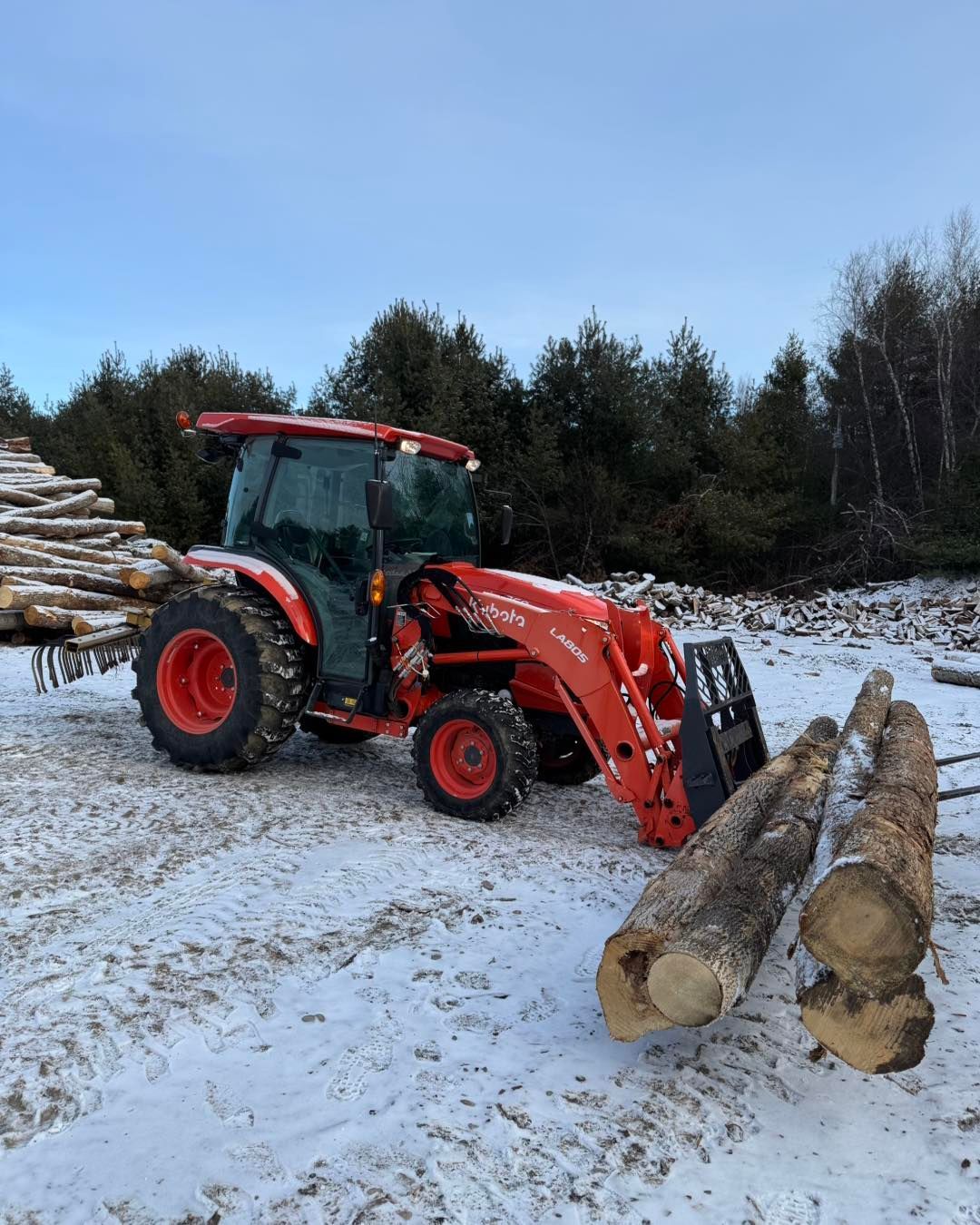 A red tractor is loading logs in a snowy field.
