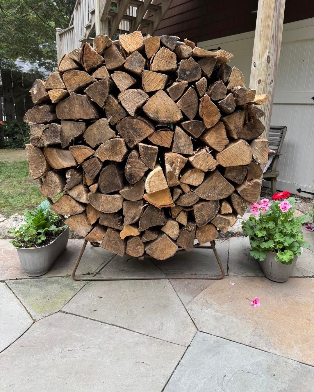 Round stack of firewood on metal stand, flanked by potted plants on a stone patio.