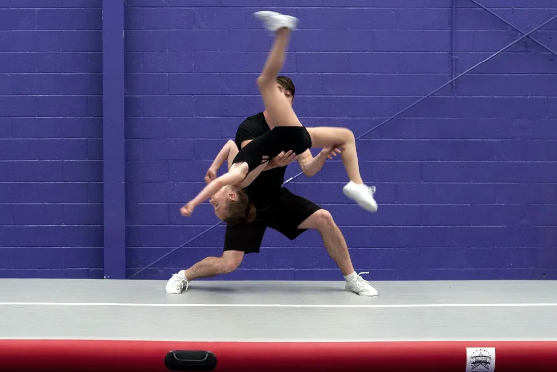 A man lifting a person doing a cheerleading move on a mat in a gym.