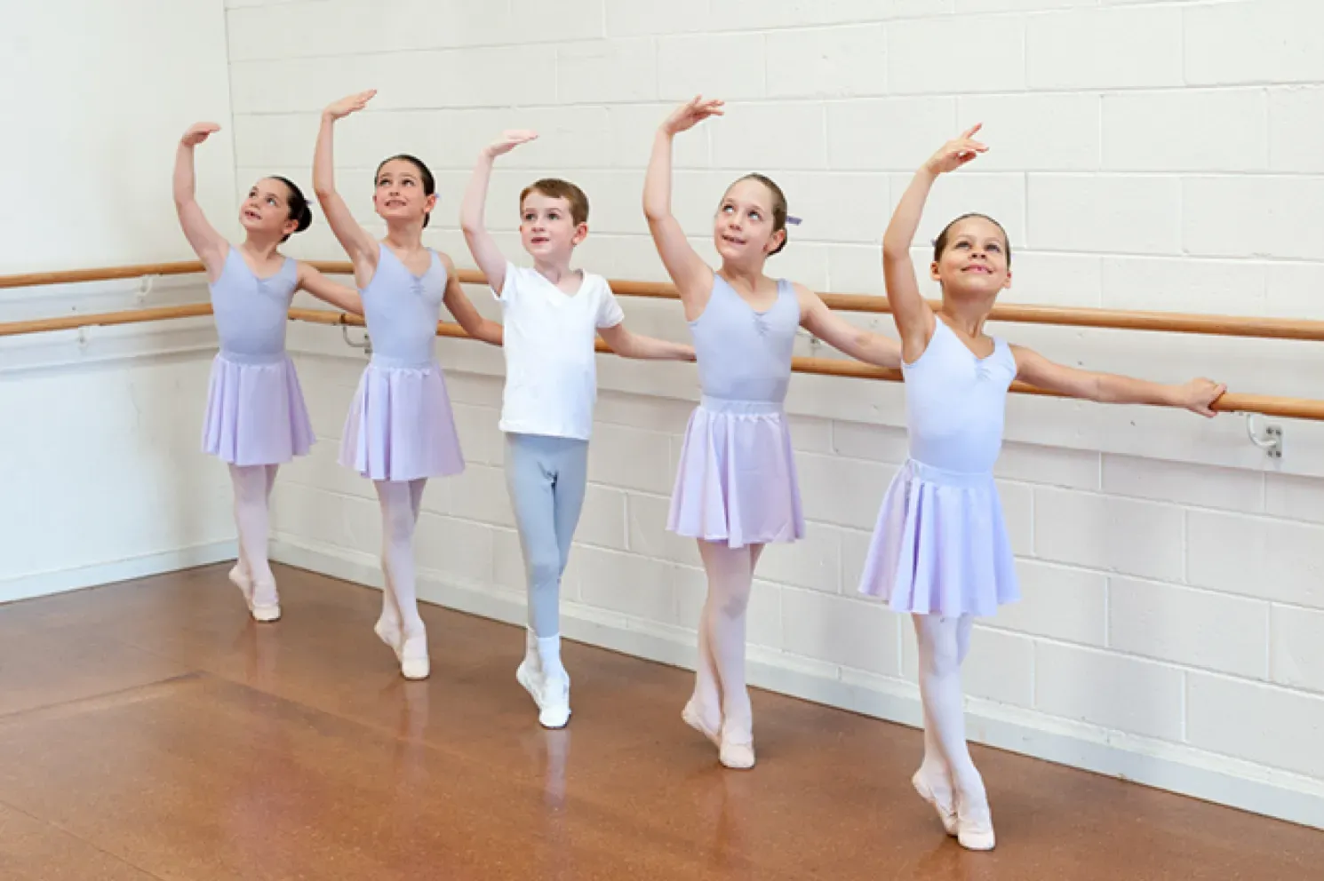 Five young ballet dancers practicing at a barre. Dancers in leotards and skirts, some with arms raised. Wooden floor and wall.