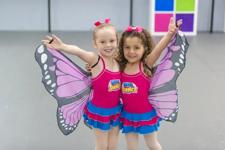 Two young girls in pink butterfly costumes smile and pose in a dance studio.