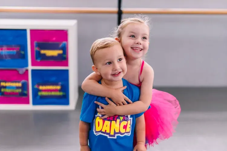 Boy and girl in a dance studio, hugging. Girl wears pink tutu. Boy in blue 