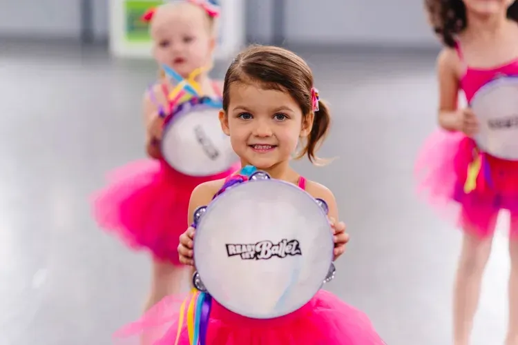 Young girl smiles, holding a tambourine, wearing a pink tutu. Other children with tambourines in background.