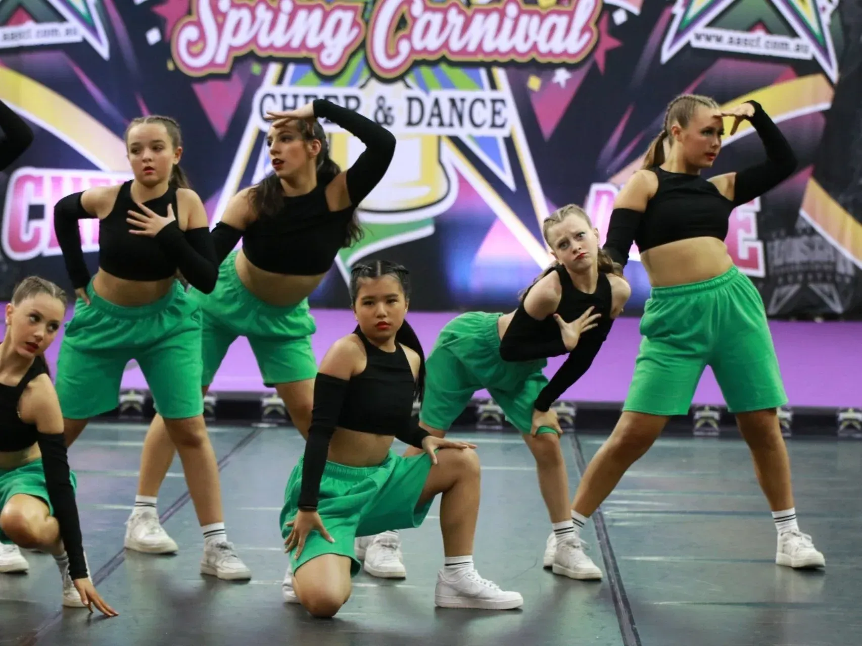 Dancers in black tops and green shorts perform a routine. Spring Carnival backdrop.