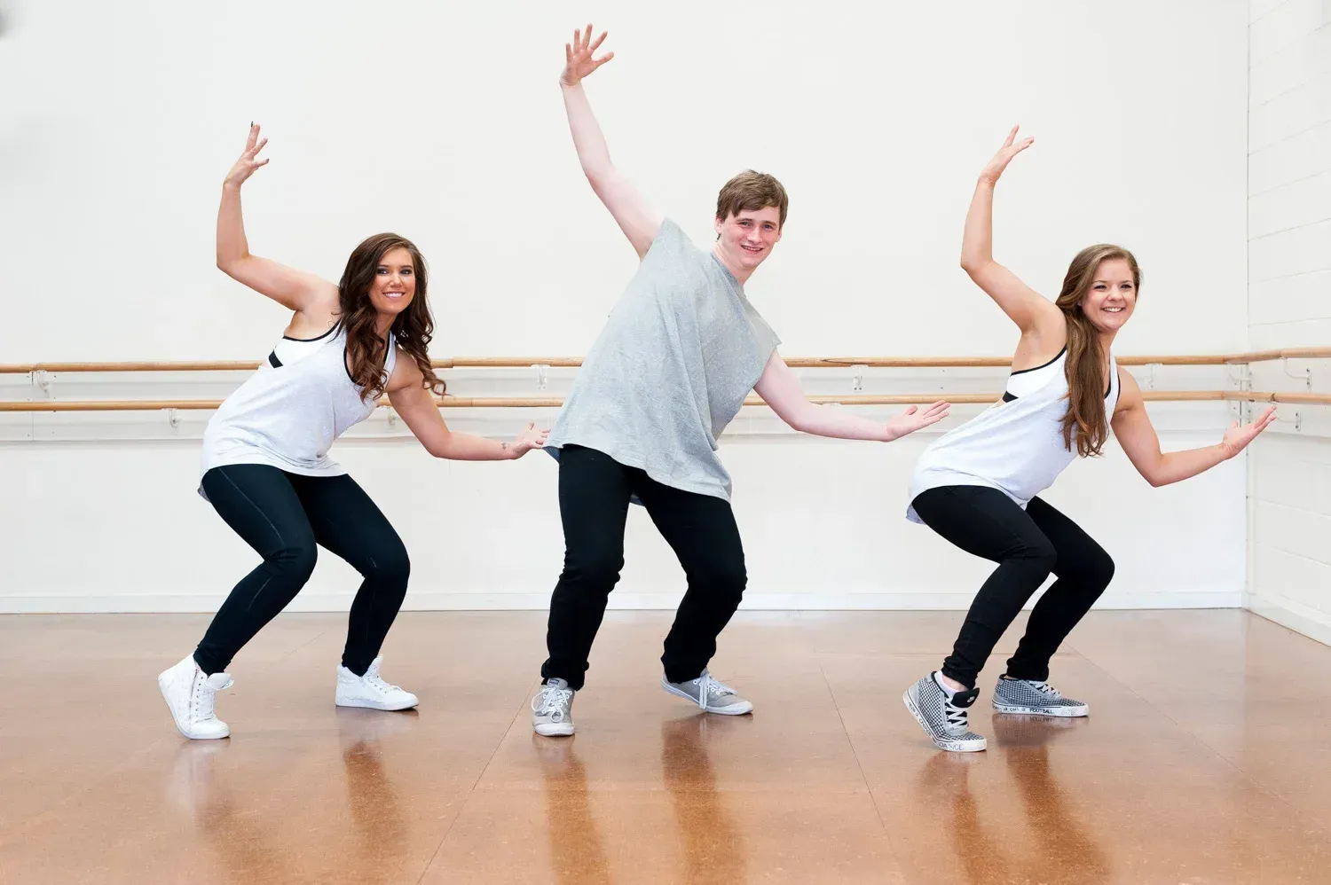 Three dancers in a studio, posing mid-movement: arms up, legs bent, smiles.