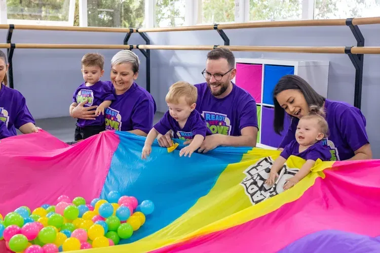 People and toddlers play with a rainbow parachute and balls, indoors.
