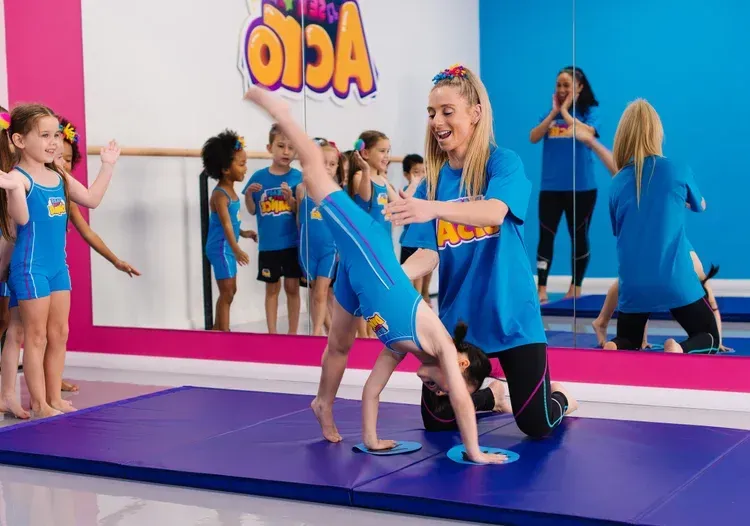 Girl doing a cartwheel with instructor's help in a gymnastics studio, children watch. Bright blue and pink decor.