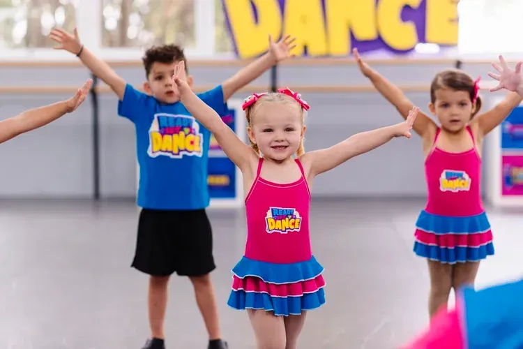 Kids dancing in a studio. One girl smiles, arms out, wearing pink; others wear blue shirts/leotards.
