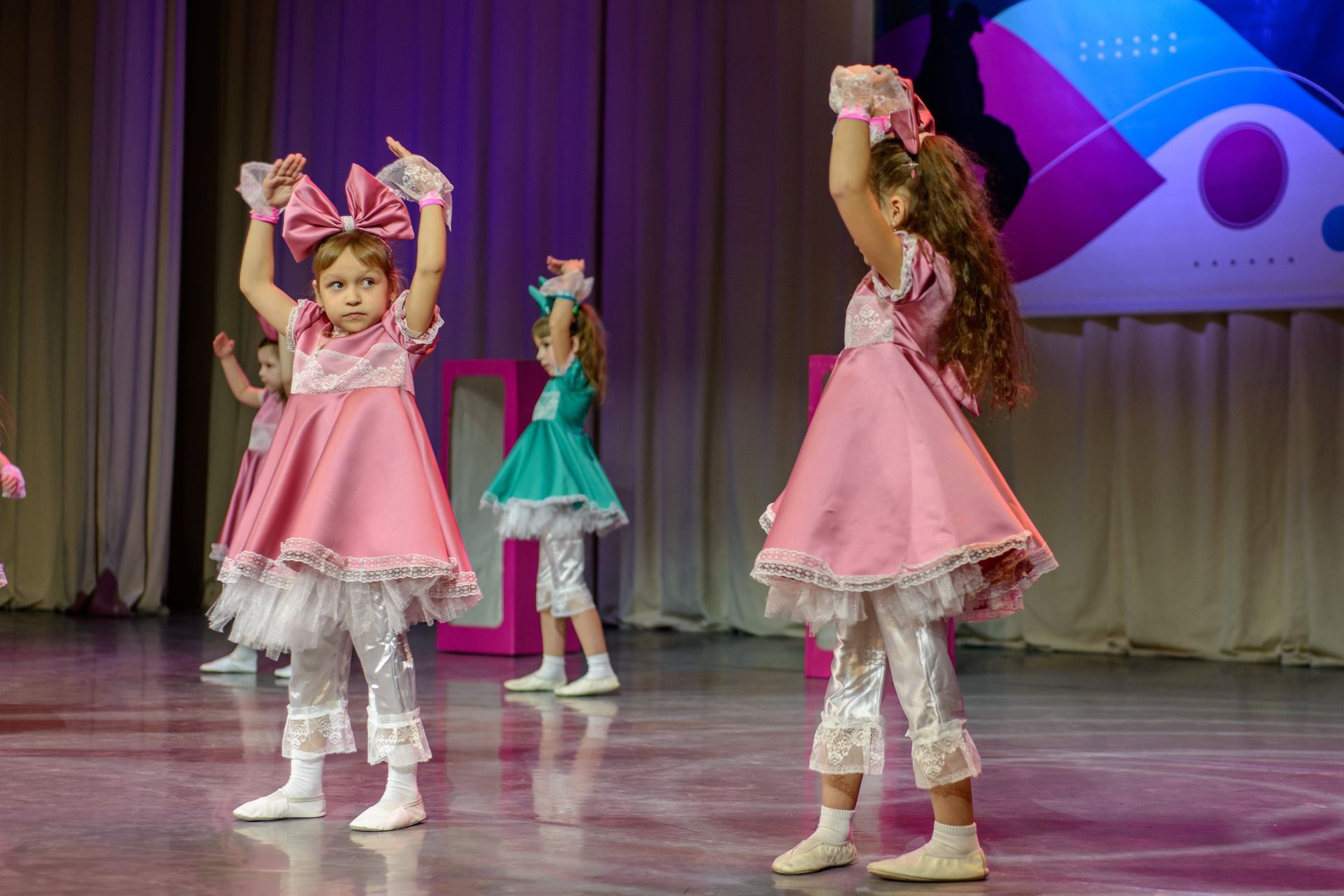 Young girls in pink and teal dresses dance on stage, arms raised, with bows and lace.