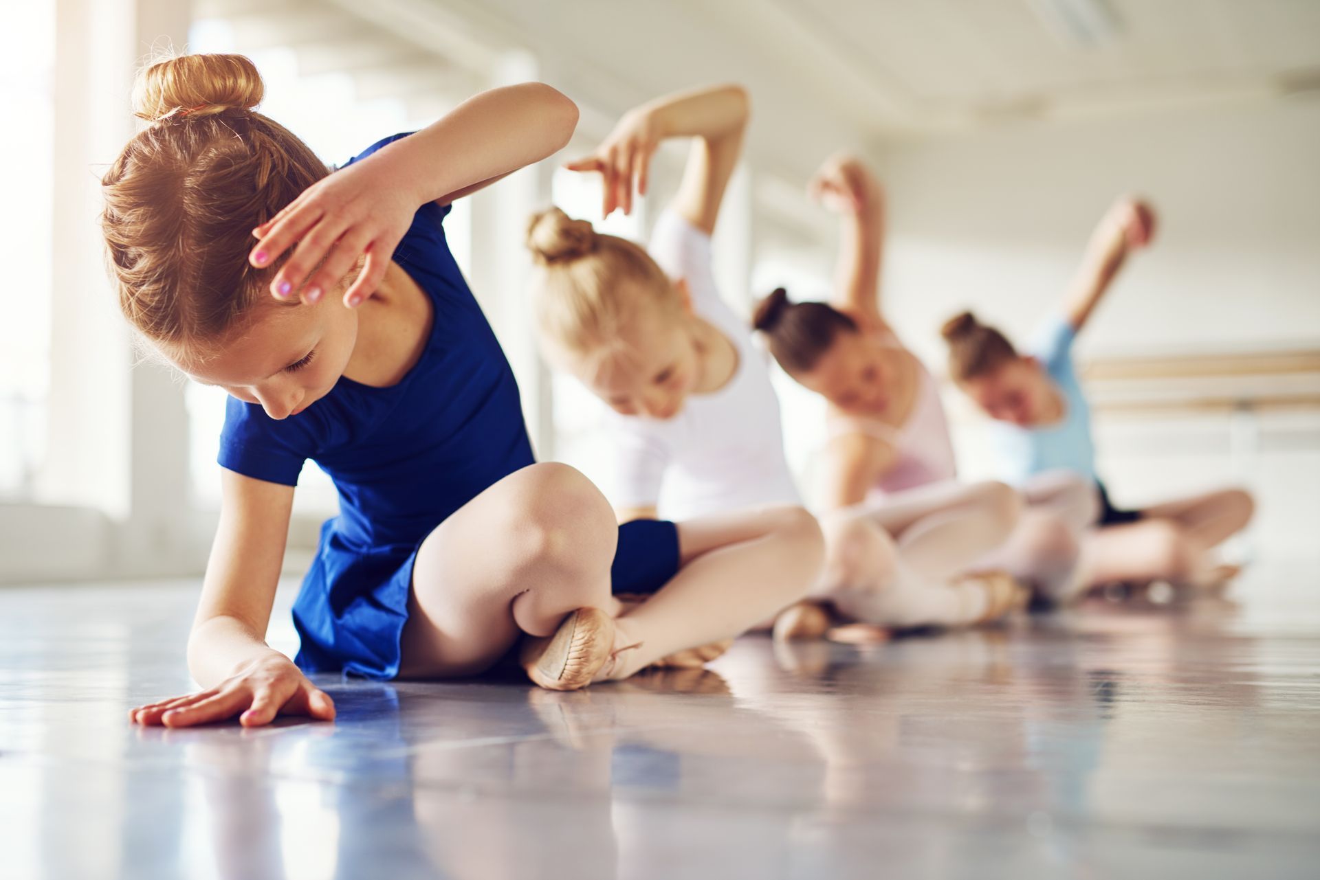 Group of young dancers in a studio, practicing a seated ballet pose with arms and hands raised.