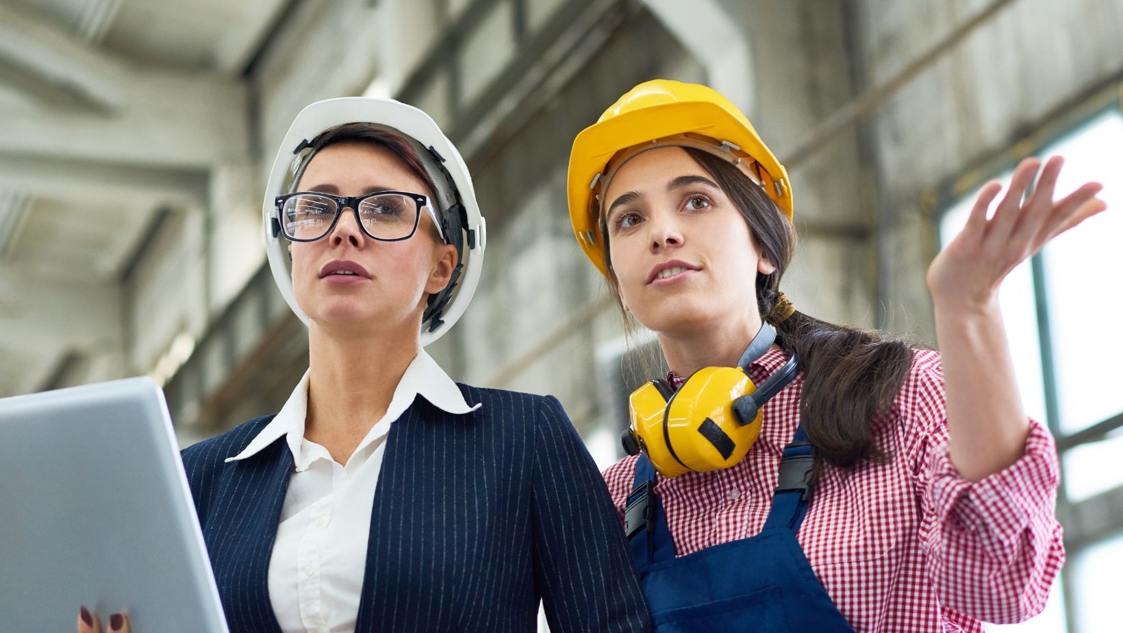 Two women in hardhats converse in a warehouse.