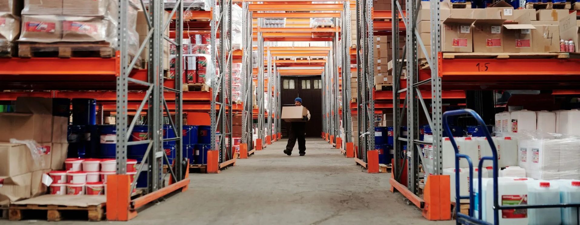This is an image of a man in a warehouse for wholesale distributors. The man is carrying a box through shelves of products.