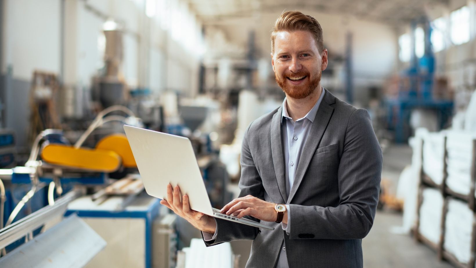 A man in a suit is holding a laptop computer in a factory.