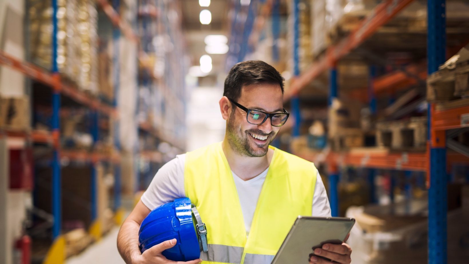 A man is holding a blue helmet and a tablet in a warehouse.