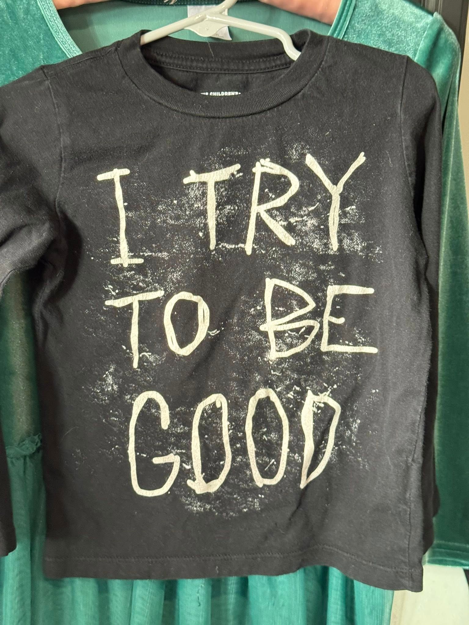 Dark gray long-sleeve shirt with the phrase “I TRY TO BE GOOD” in white lettering on a hanger.