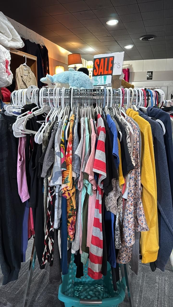 Clothing racks in a thrift store, with a sale sign hanging above colorful shirts and dresses.