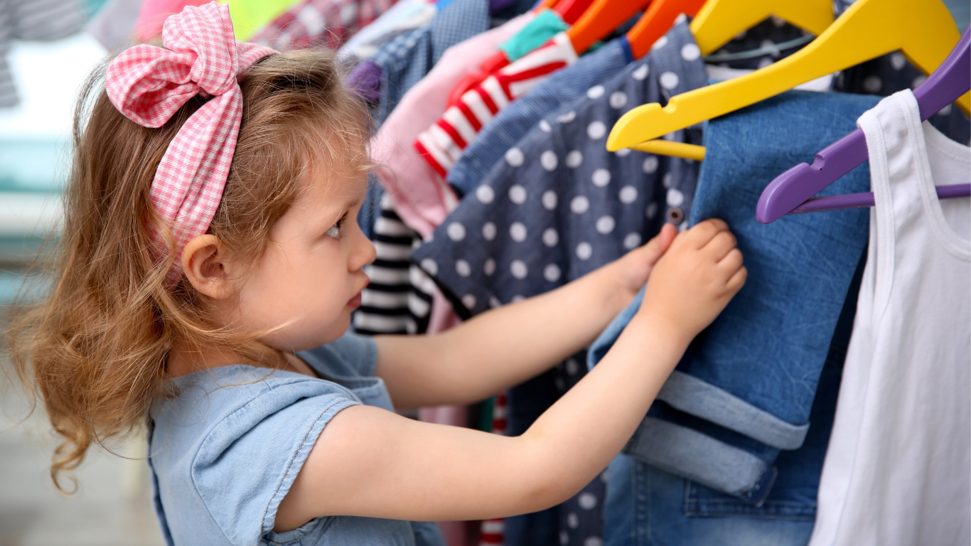 Child hanging clothes on hangers in a closet, reaching toward blue denim and striped shirts.
