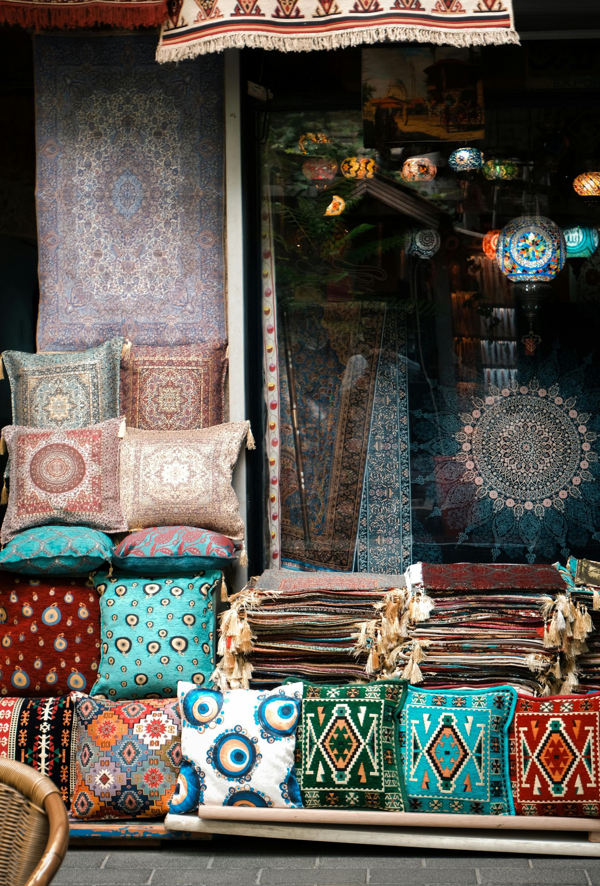 Colorful patterned textiles and cushions displayed in a shop window, with folded rugs and hanging lanterns.