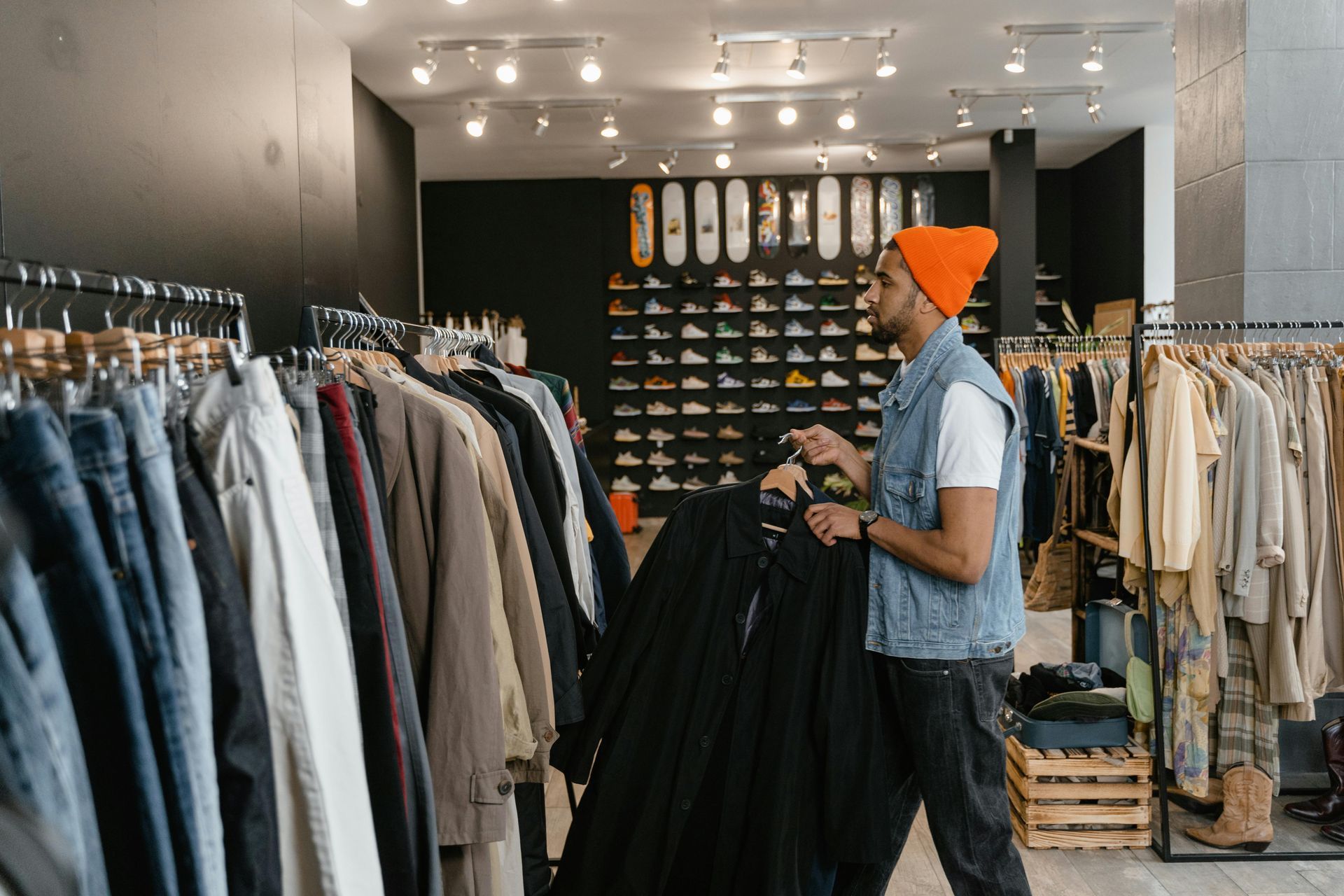 Person browsing clothes in a boutique, wearing an orange beanie and denim vest.