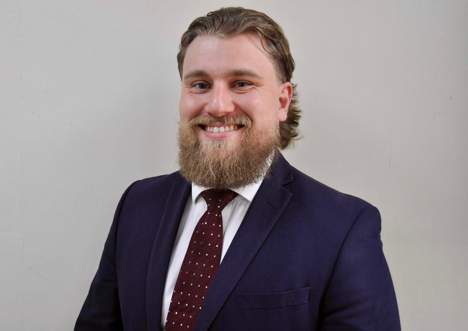 A person with a beard, wearing a navy suit, white shirt, and patterned maroon tie, smiling against a neutral background.