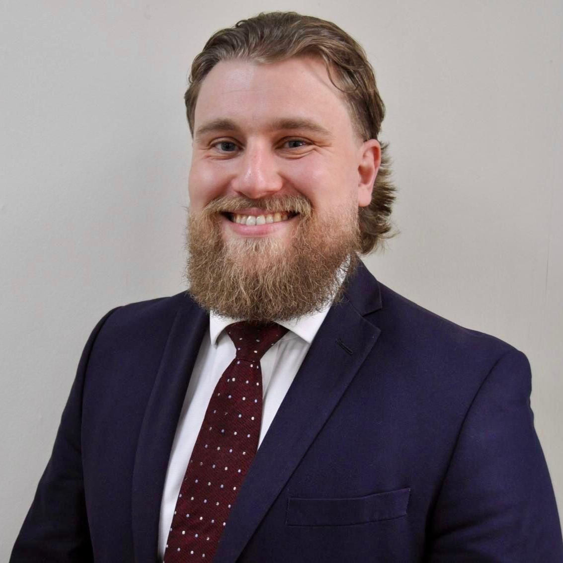 A person with a beard and hair pulled back, wearing a navy blue blazer, white shirt, and dark patterned tie.