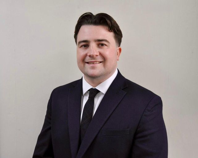 A person in a dark suit, white shirt, and black tie posing against a light gray background.