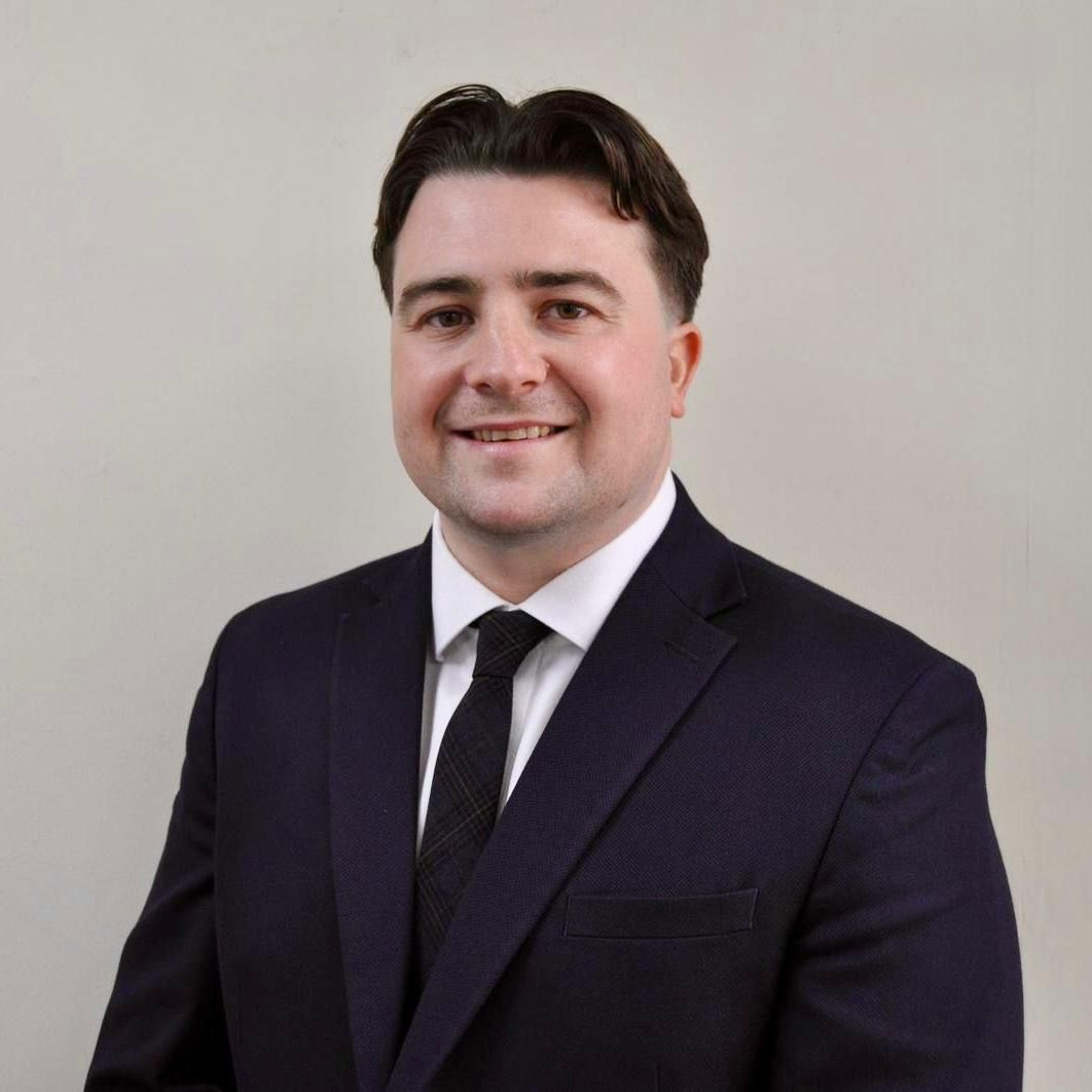 A smiling person with dark hair, wearing a navy suit, white collared shirt, and black tie against a light gray background.