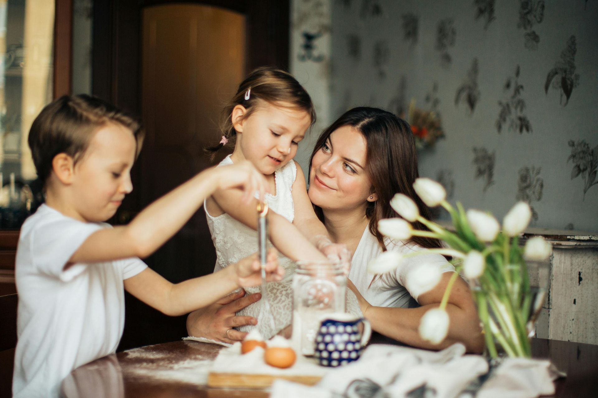 Two children and a woman share a candlelit craft table with flowers and supplies