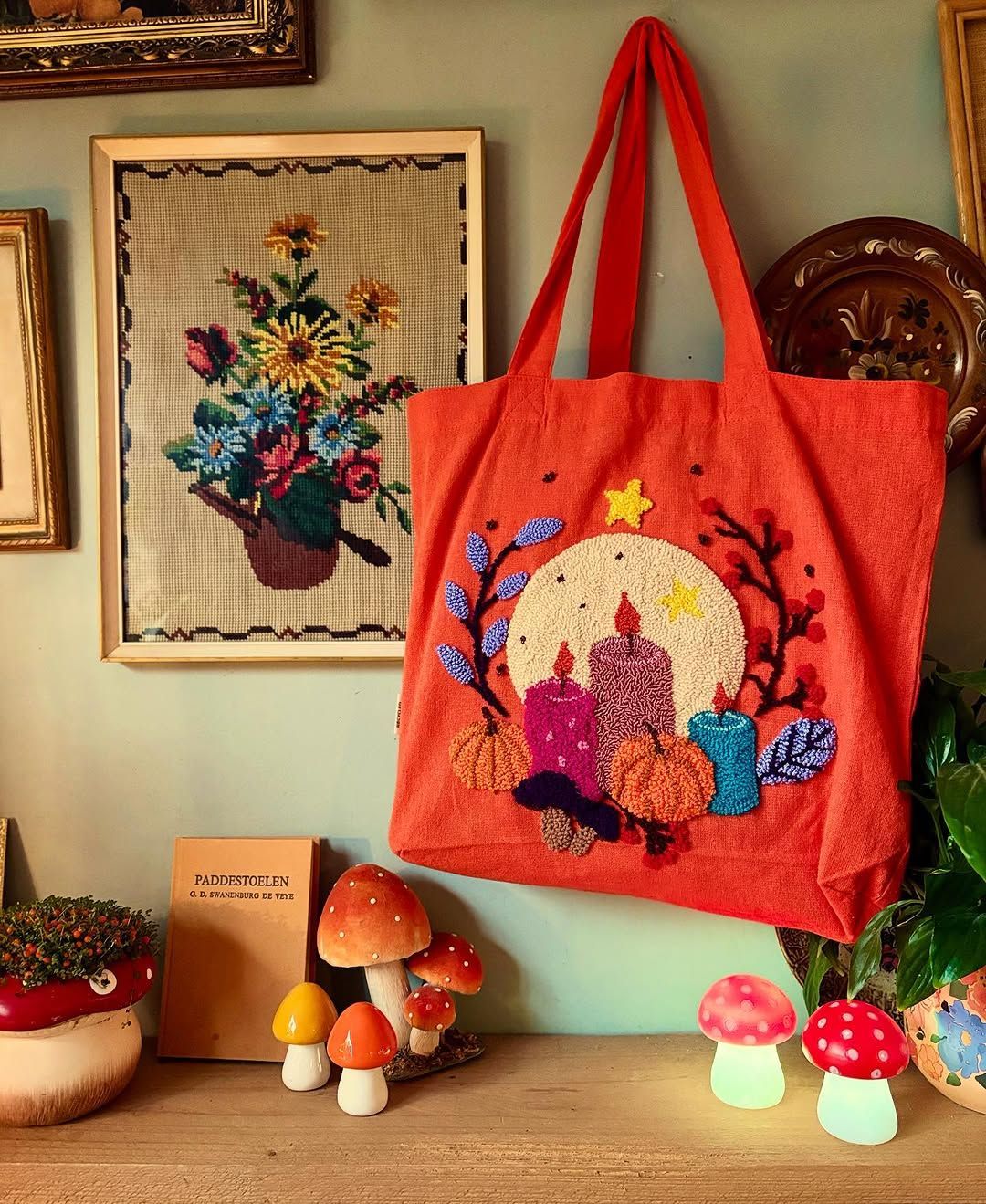 Een oranje draagtas met geborduurde kaarsen hangt aan de muur naast ingelijste kunst en paddenstoelbeeldjes.