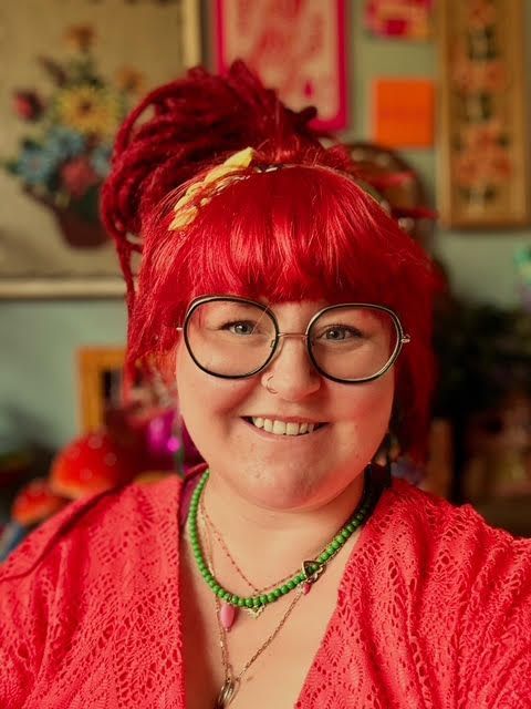 Een vrouw met rood haar en een bril glimlacht. Ze draagt ​​een rode top en kralenkettingen, tegen een kleurrijke achtergrond.