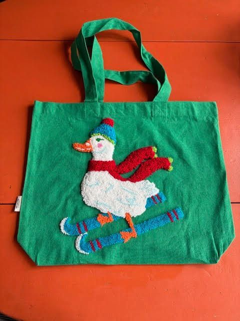 Groene draagtas met een witte skiënde eend, die een rode sjaal en een blauwe muts draagt.