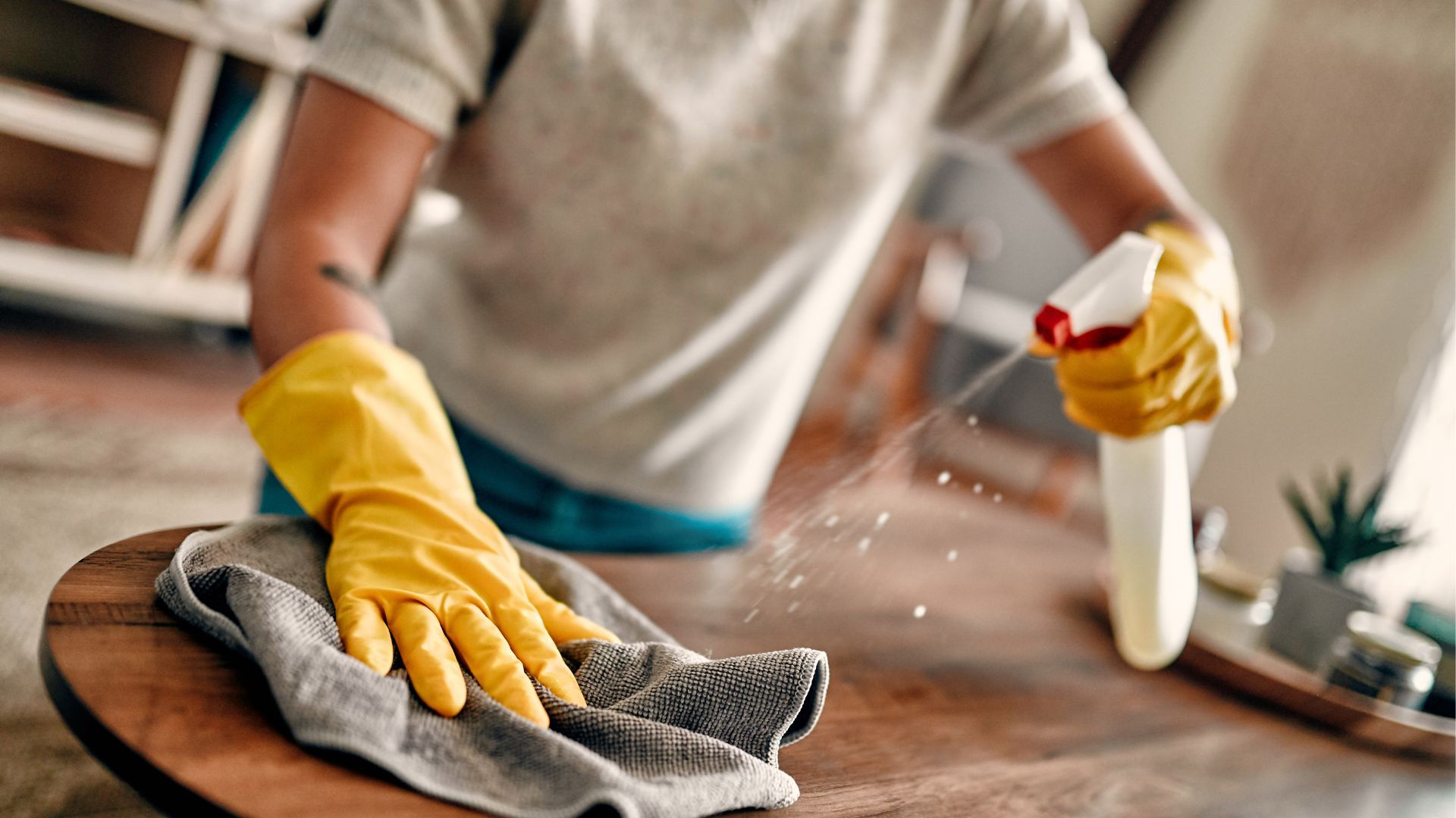 Person wearing yellow gloves sprays and wipes a wooden table with a cloth.