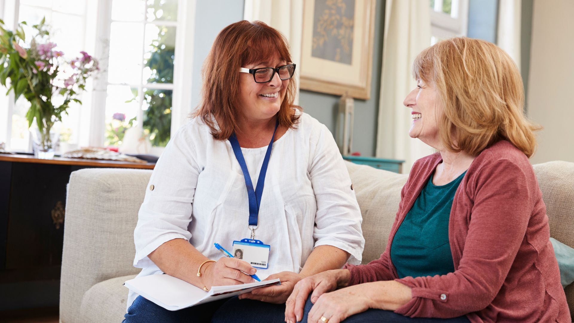 Woman in glasses taking notes while talking to another woman on a couch. Living room setting.