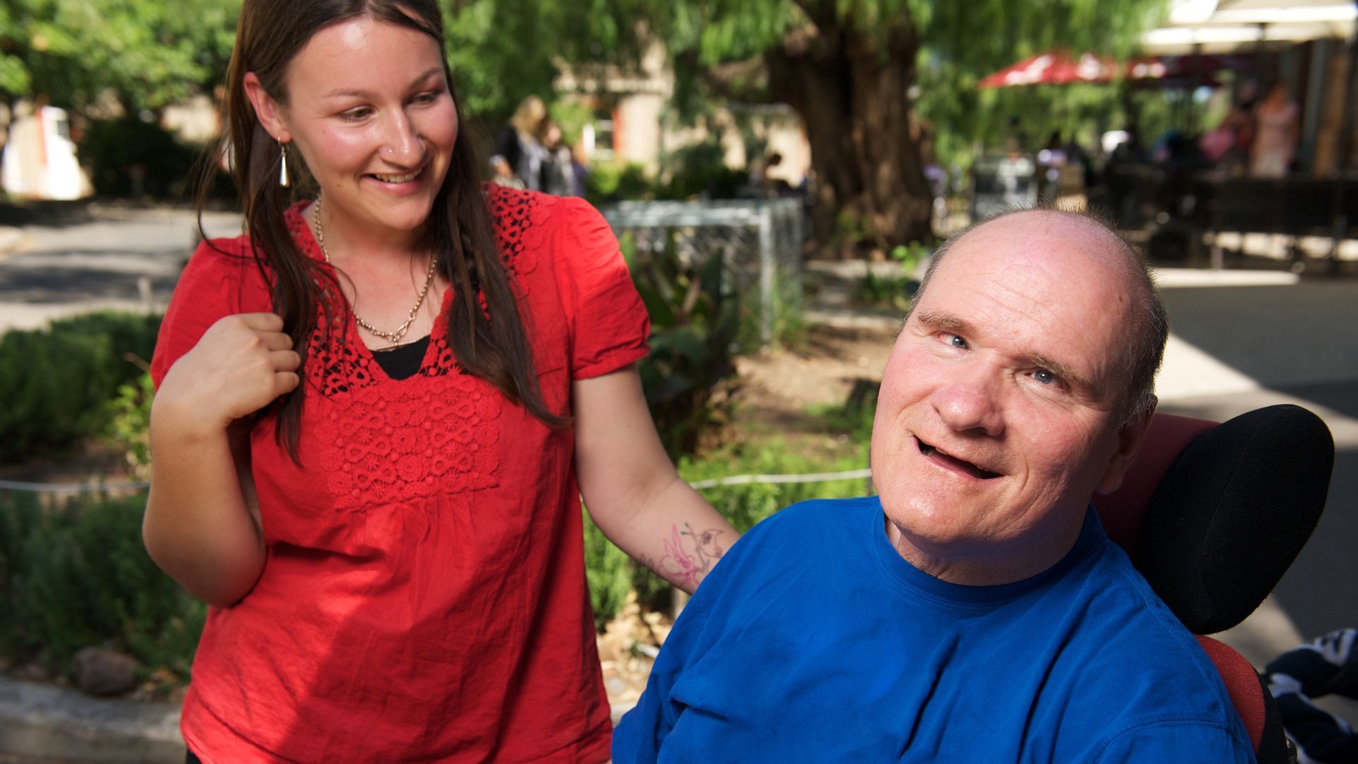 Woman smiles, touches the shoulder of a man in a wheelchair; they're outside in the sun.