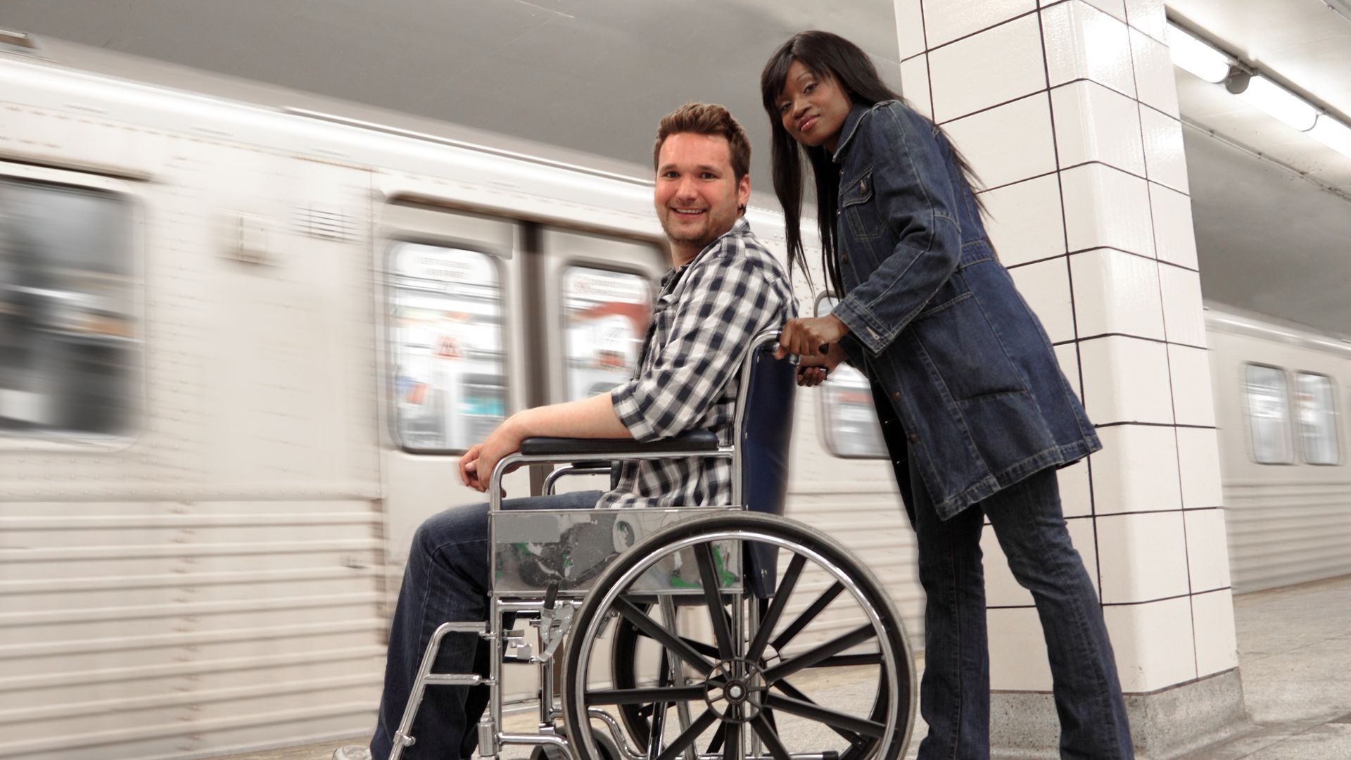 Woman pushing a person in a wheelchair toward a subway train. They're smiling in a station.