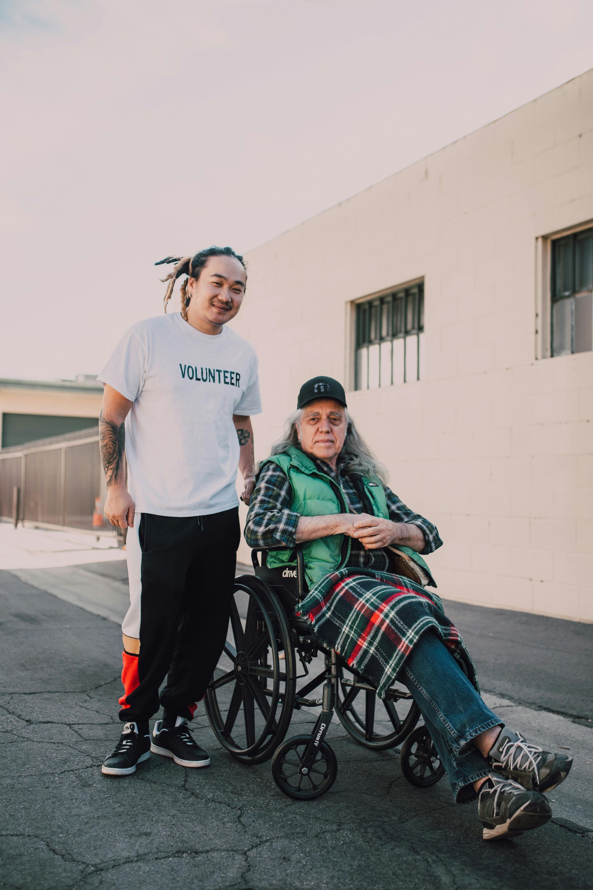 Person in wheelchair with a companion, outdoors. Brightly lit street. Wheelchair user wearing a plaid coat, companion in a white shirt.