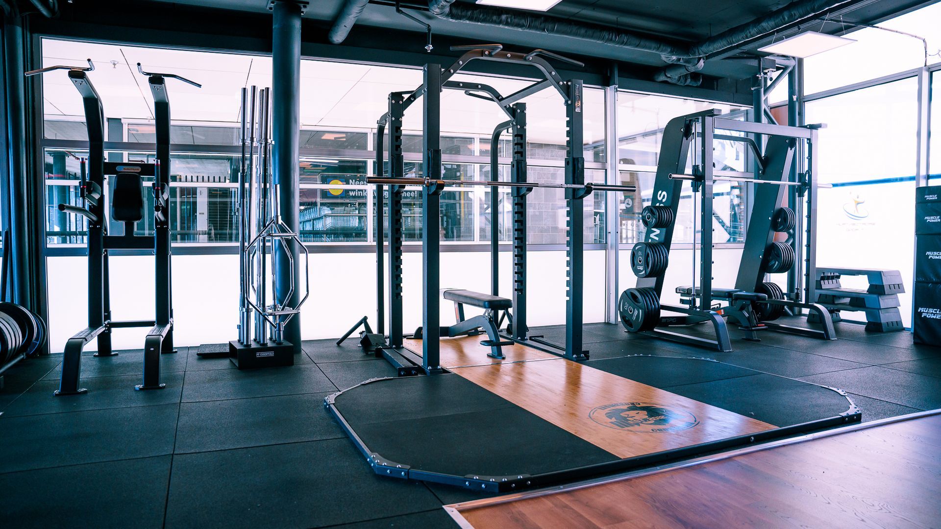 Interieur van een fitnessruimte met fitnessapparatuur bij een raam. Voorzien van een power rack, bank, optrekstangen.