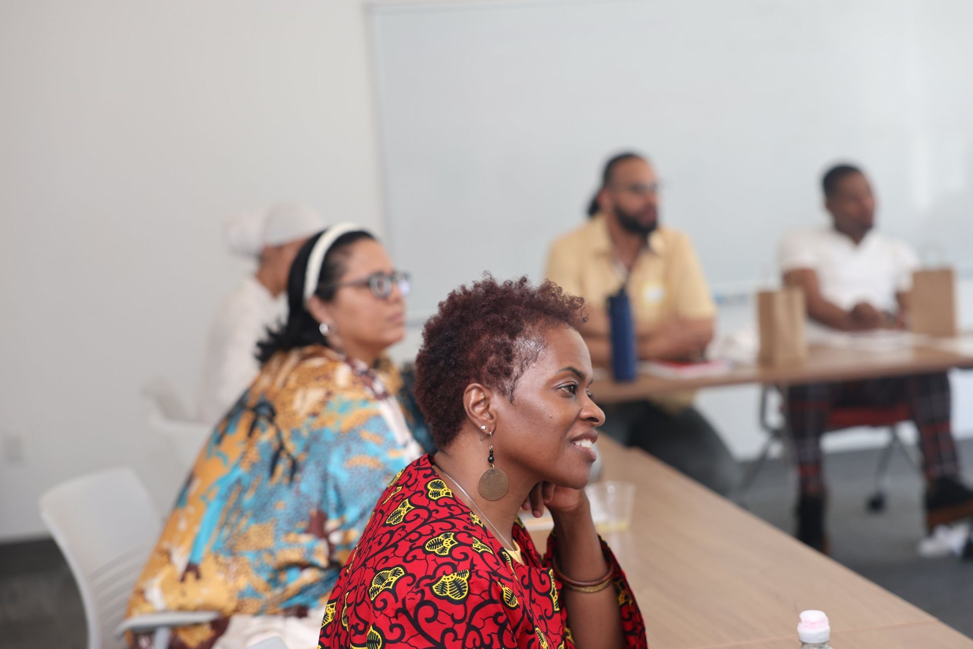 Attendees at a meeting. Woman in red patterned shirt smiles. Others sit at a long table.