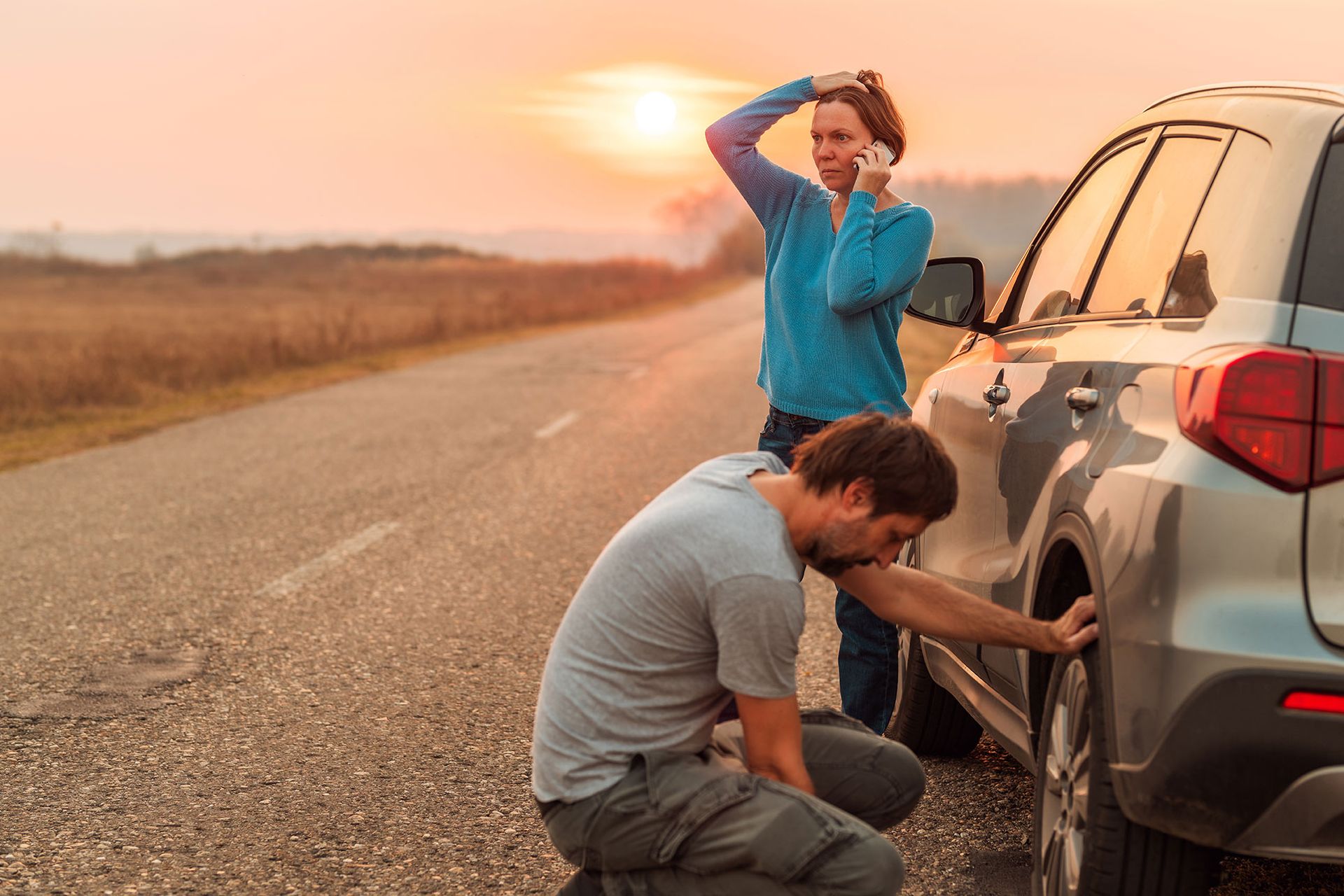 Man changing a flat tire on a car as a woman talks on the phone on a rural road at sunset.