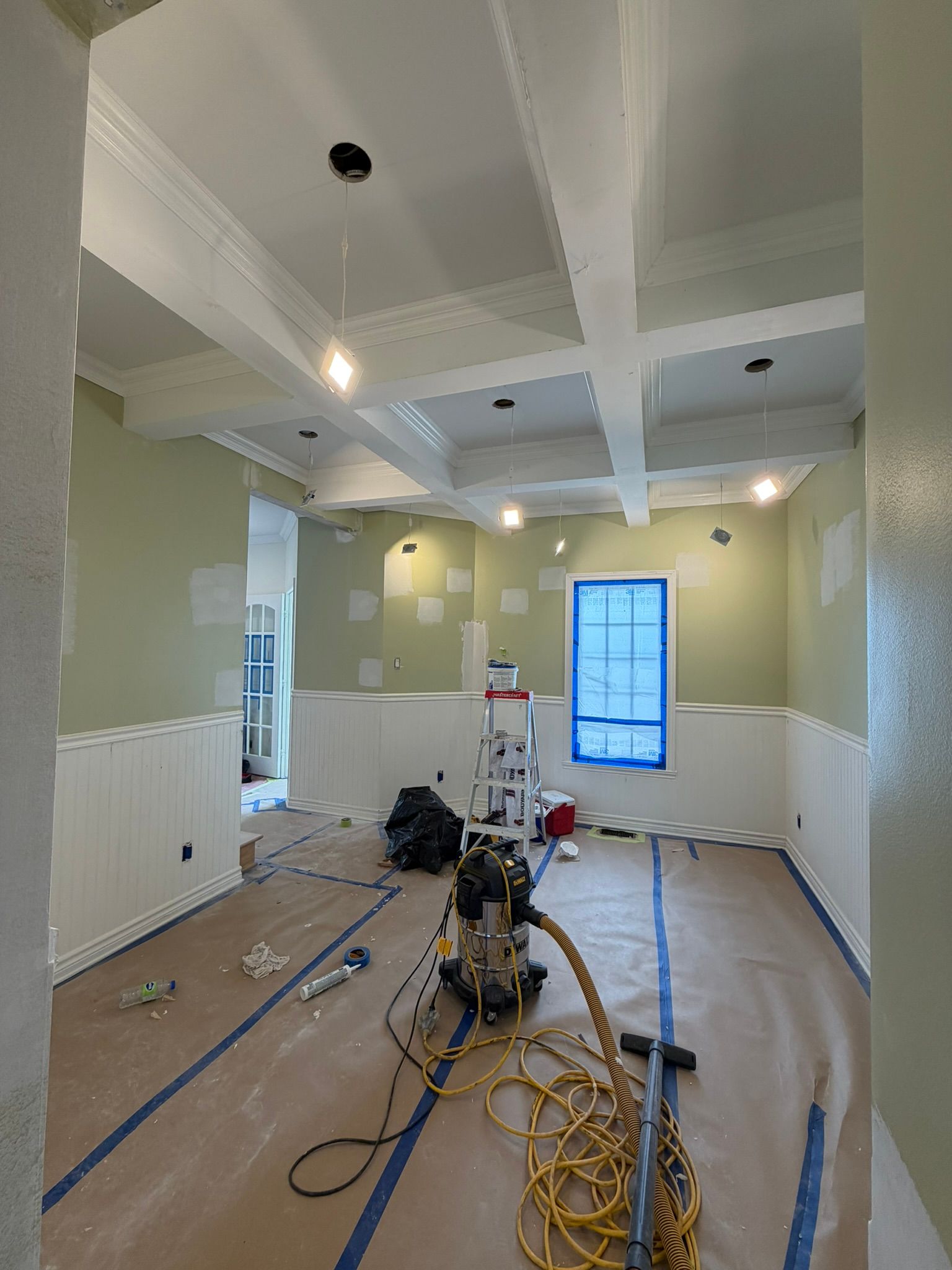 Pièce en rénovation avec poutres blanches au plafond, murs vert pâle, outils, câbles et une fenêtre recouverte de ruban adhésif bleu.