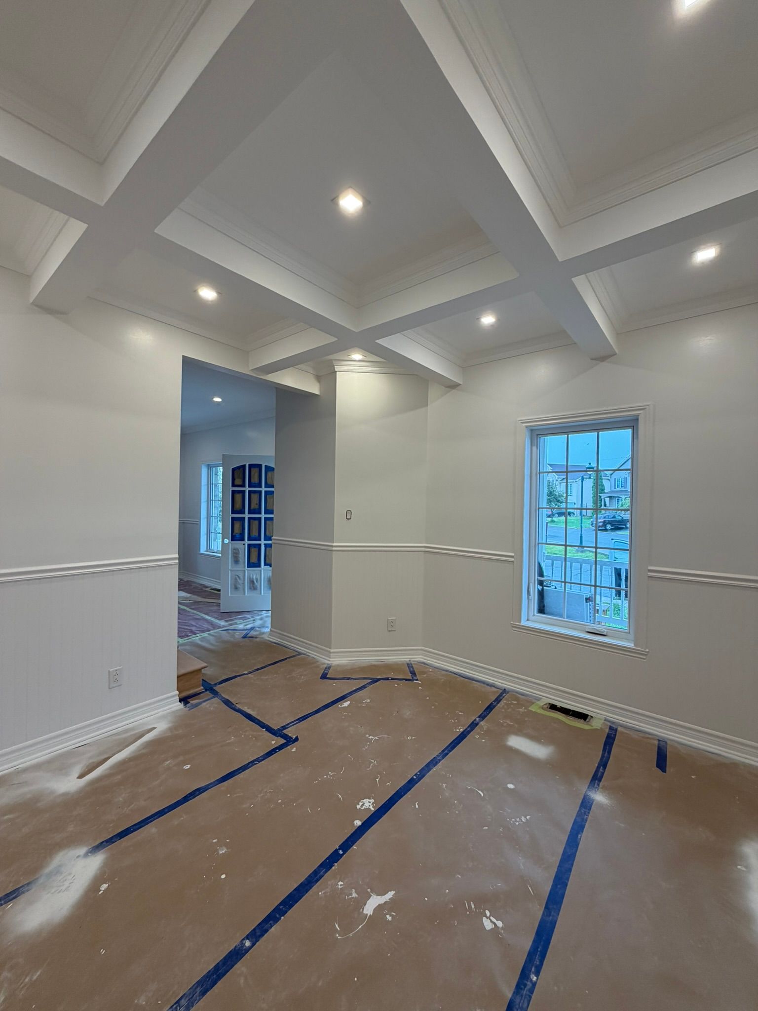 Pièce vide en rénovation, avec plafond à caissons blanc, spots encastrés, sol recouvert de ruban adhésif et deux fenêtres.