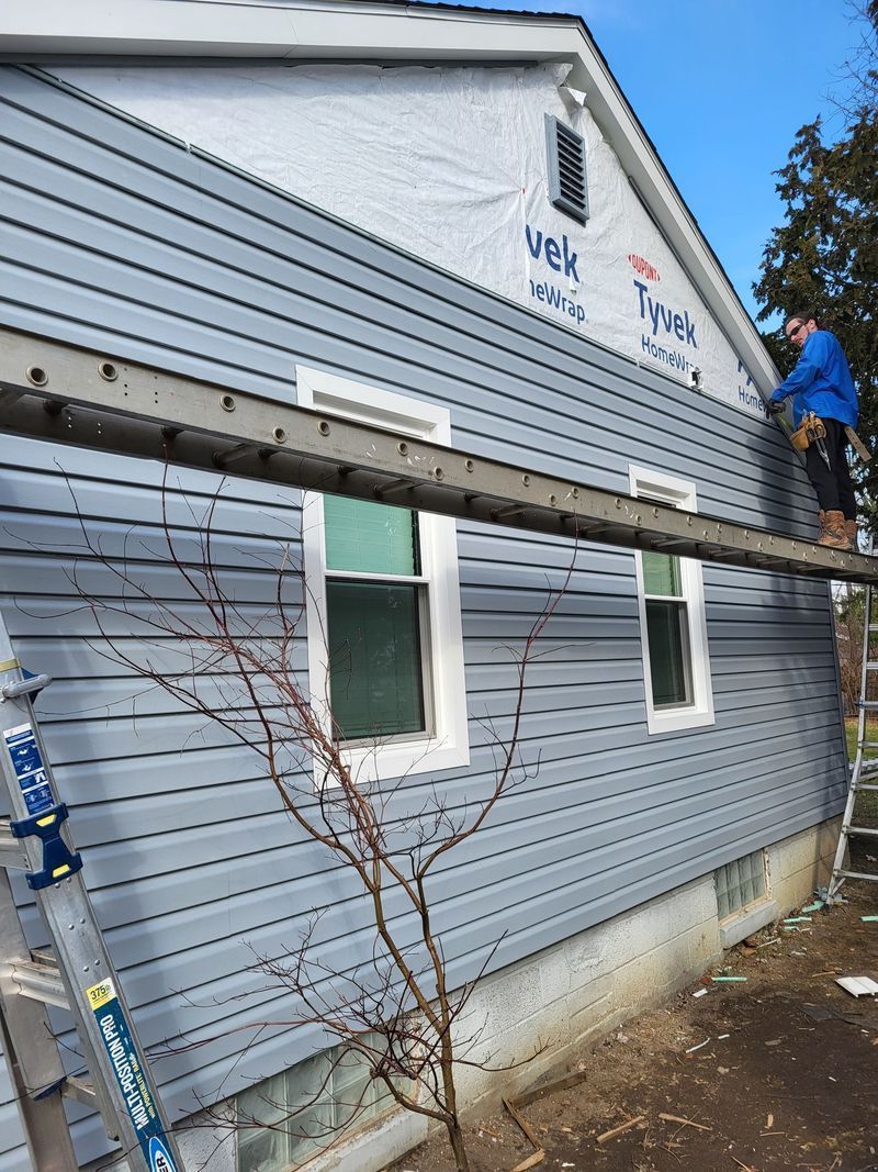A person installing blue siding on a house, standing on a ladder. The house has white windows and Tyvek wrap.