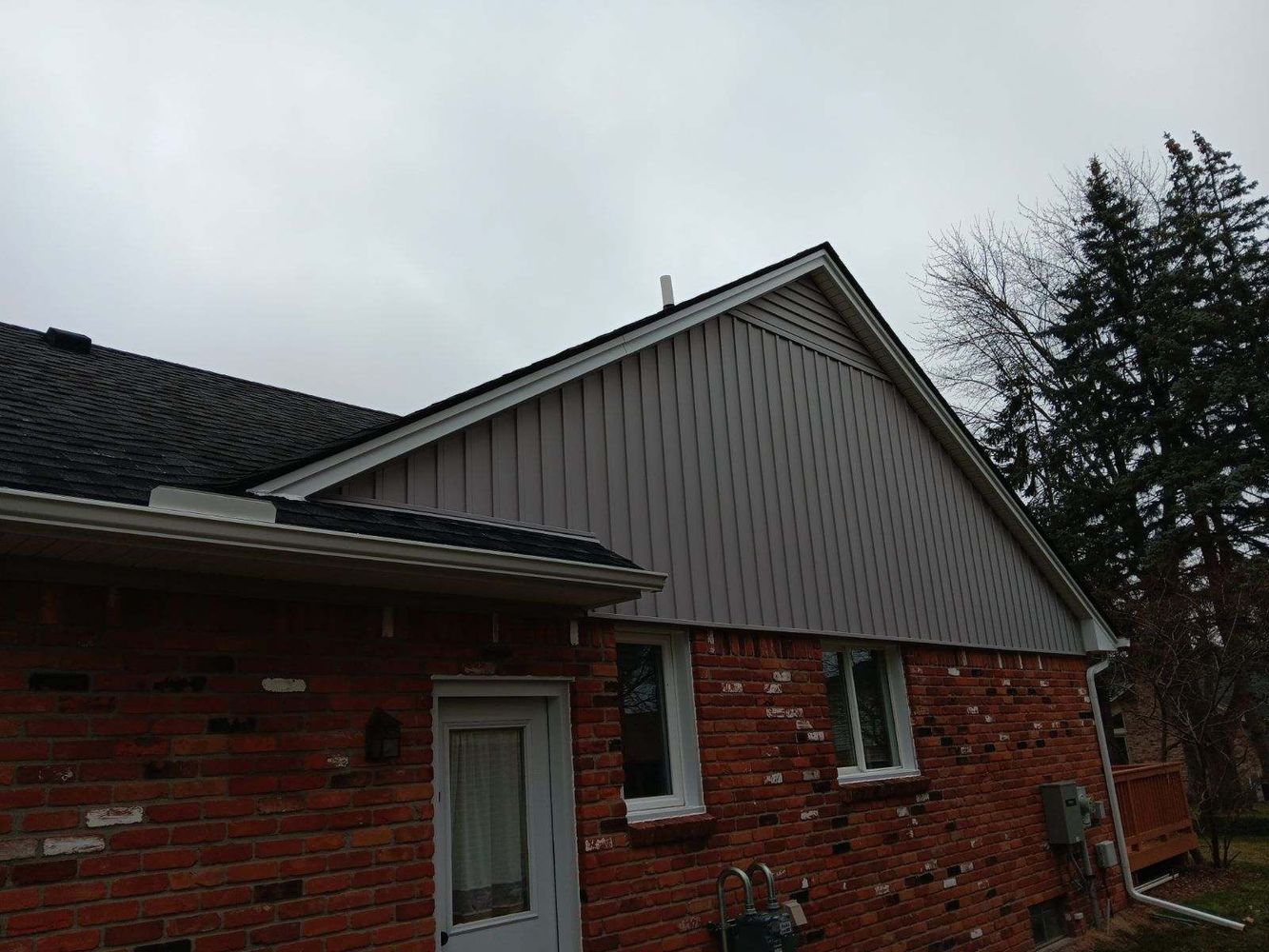 Brick house exterior with gray siding, black roof, and overcast sky.