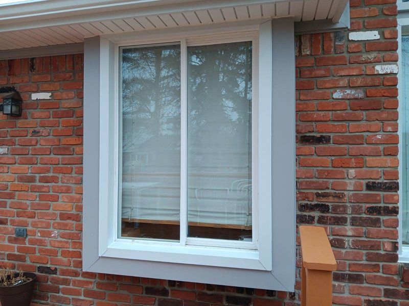 A rectangular window set in a brick wall. The window has a gray frame and white blinds.