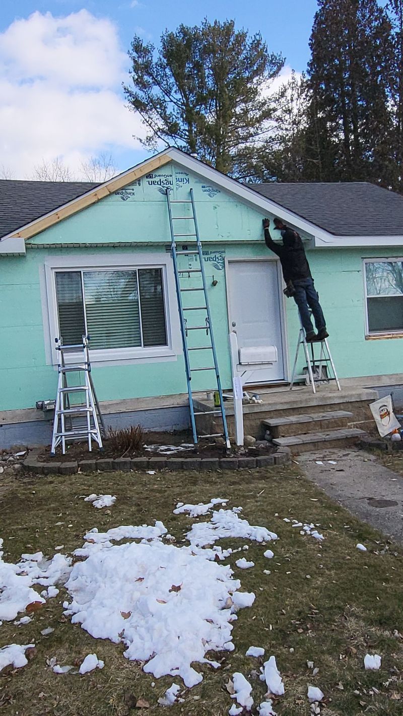 Person on ladders working on a house exterior. Green siding, snow on the ground, blue sky.