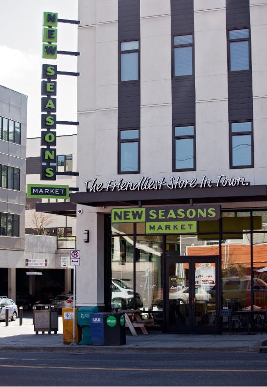 New Seasons Market storefront on a city street, with vertical sign and glass entrance.