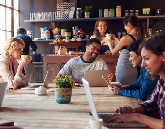 People working and chatting in a cozy café with laptops and drinks at a wooden table