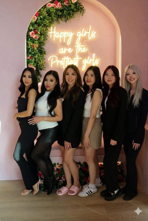 Six women posing in black and white outfits before a floral arch with a neon sign reading “Happy girls are the prettiest girls.”