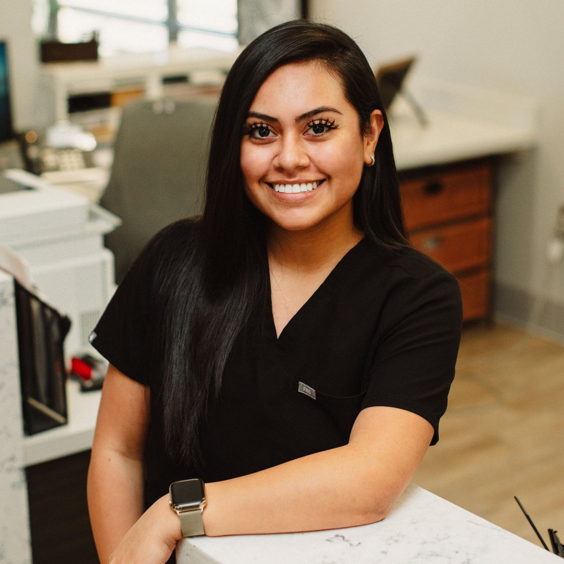 A woman in a black scrub top is smiling while sitting at a desk.